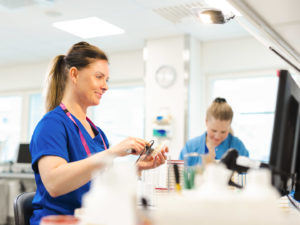 Naispuolinen työntekijä pitelee käsissään näytettä ja katsoo tietokoneen näyttöä. Taustalla näkyy toinen naispuolinen työntekijä.